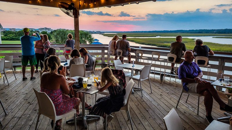 people sitting on tables on deck of Jinks Creek Waterfront Grill watching a pink sky sunset over the salt marshes