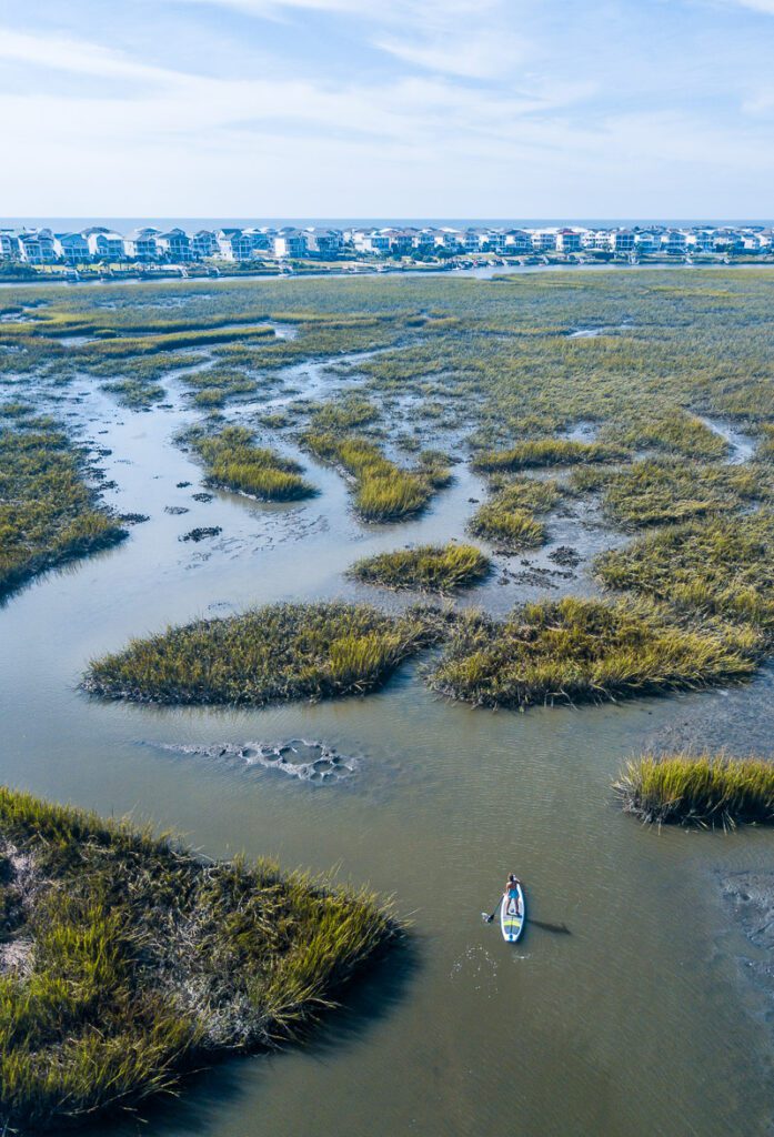 aerial view of women Stand up paddle boarding the salt marshes with buildings and beach in the background