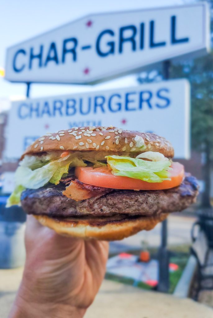 Burger with lettuce, tomato, in front of the Char Grill sign