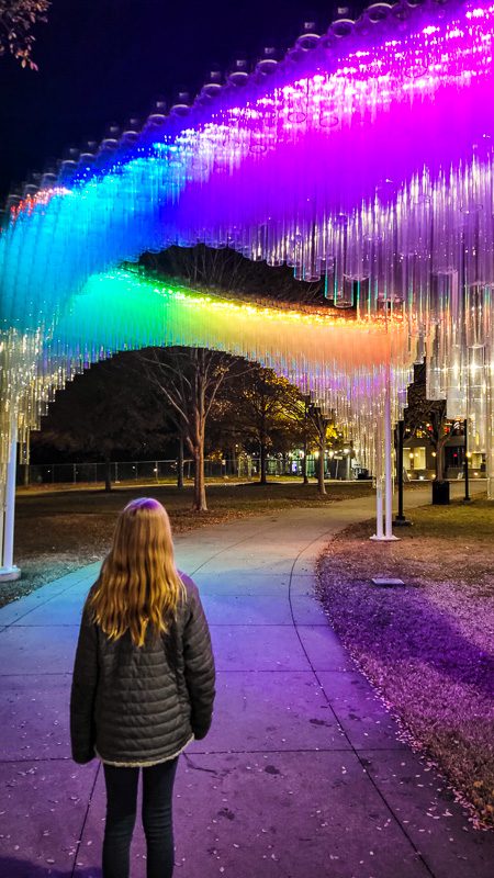 girl standing in front of rainbow colored plexi glass sculpture