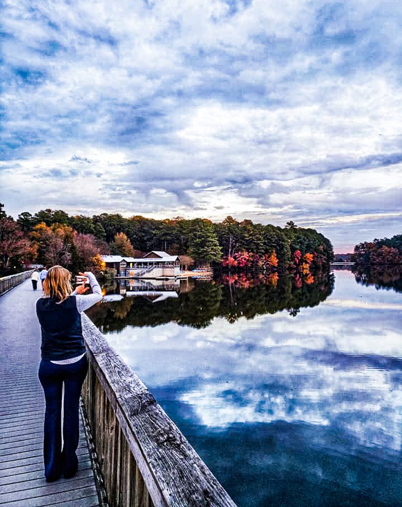 Lady standing on a bridge taking a photo of a lake