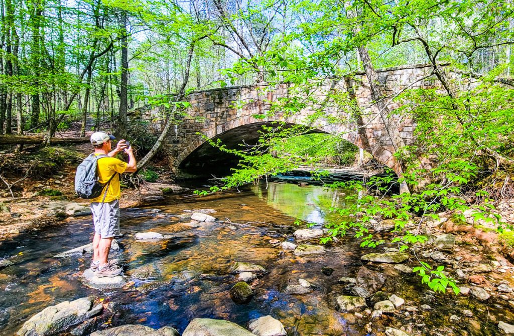 Man standing on rocks in a stream photographing a bridge