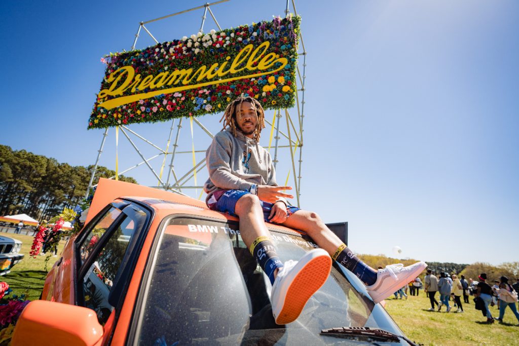 Man sitting on top of a car with a sign behind him that says Dreamville