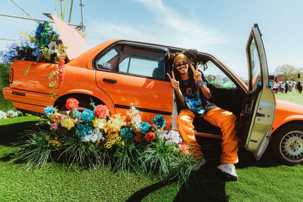 Man sitting in an orange colored car with door open and surrounded by flowers