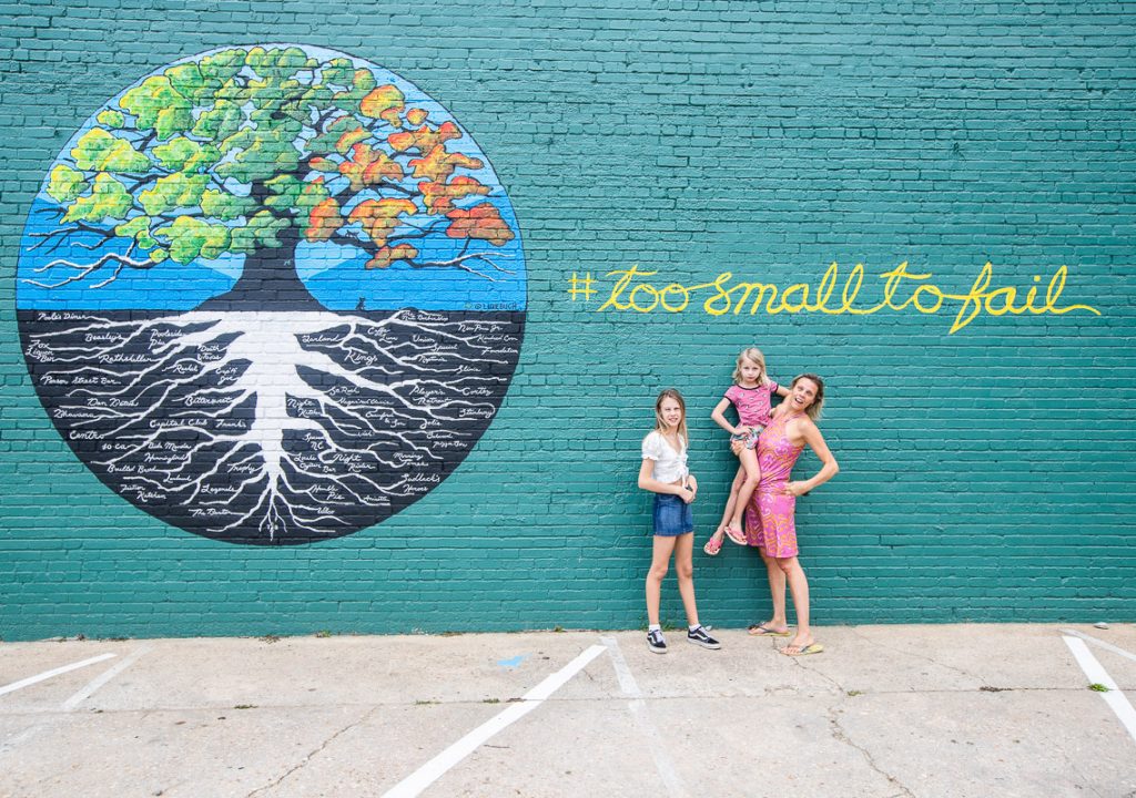 Mom and two daughters standing next to a mural that says "too small to fail"