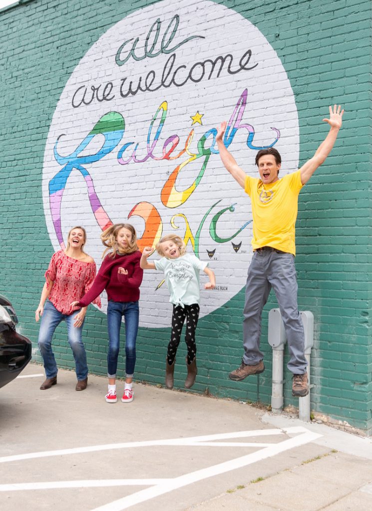 Family jumping up for a photo in front of a mural which says "all are welcome, Raleigh".
