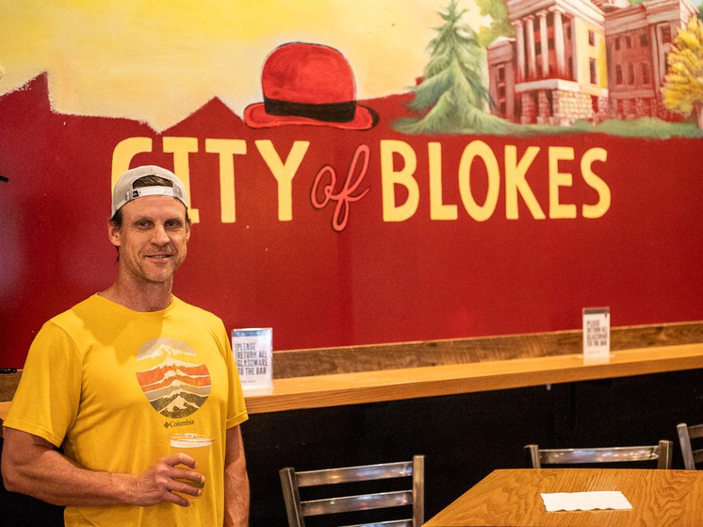 Craig Makepeace (a male) standing with a beer in his hand with the sign City of Blokes behind him