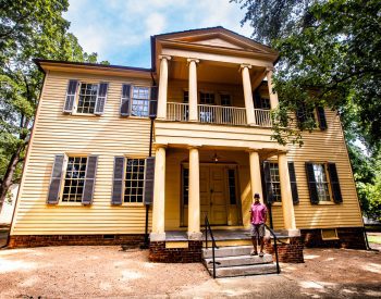 Man in a pink shirt standing on steps in front of a historic yellow house
