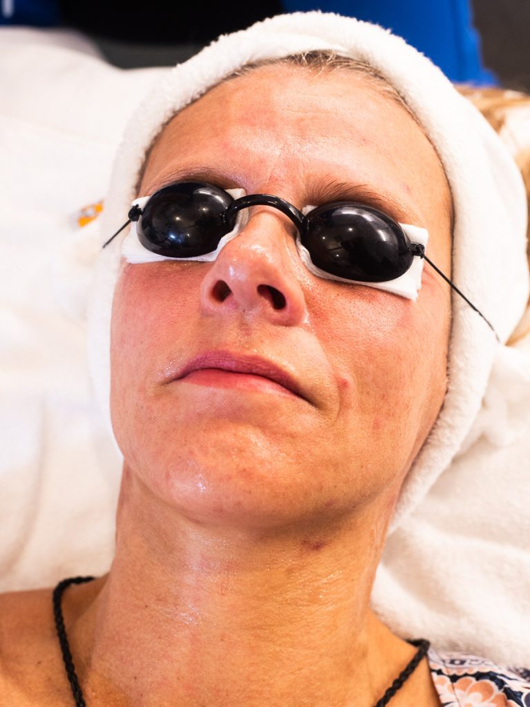 A woman lying down with black goggles over her eyes and a white towel over her hair.