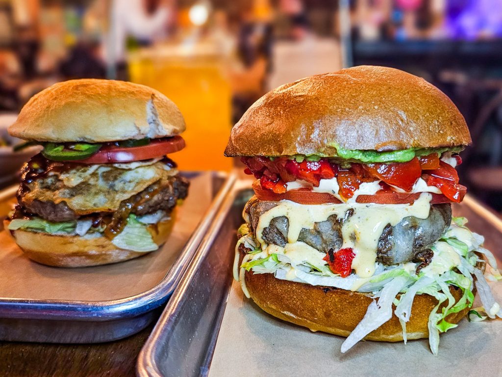Two burgers with cheese and red peppers and lettuce on trays in a restaurant.