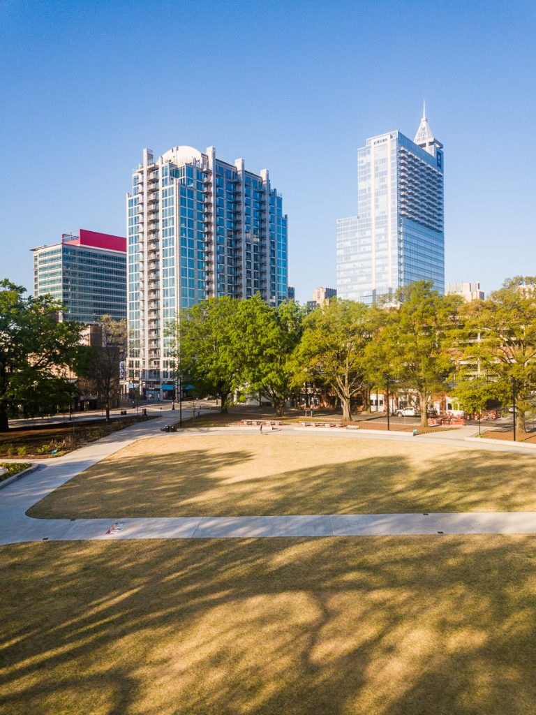 A grass area of a city park with trees and buildings in the background.