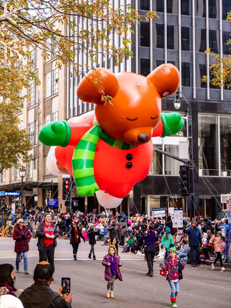A Christmas parade in a city street with a giant floating bear dressed in red and green.