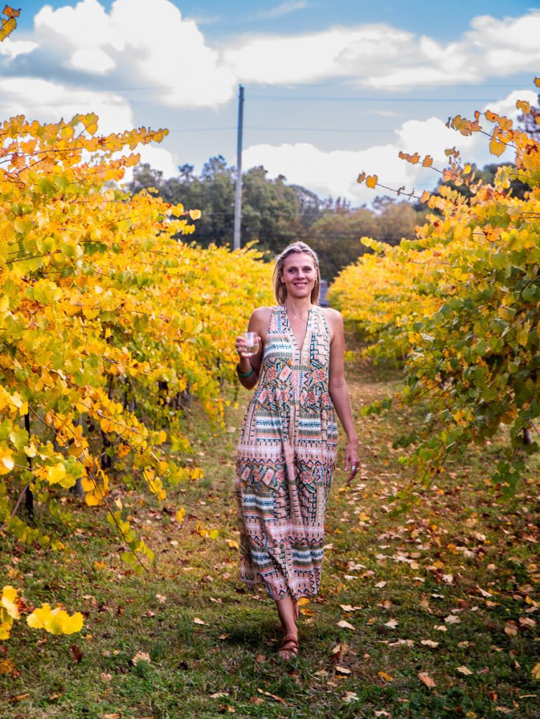 A lady walking in a vineyard drinking wine.