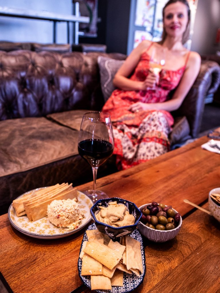 Lady sitting on a brown leather couch drinking wine with olives and chesses on the table.