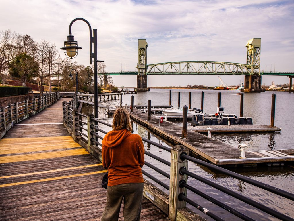 Lady walking along a riverfront boardwalk with a bridge in the distance.