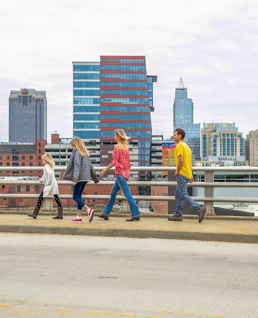 Family of four walking across a bridge with the city of Raleigh in the background.